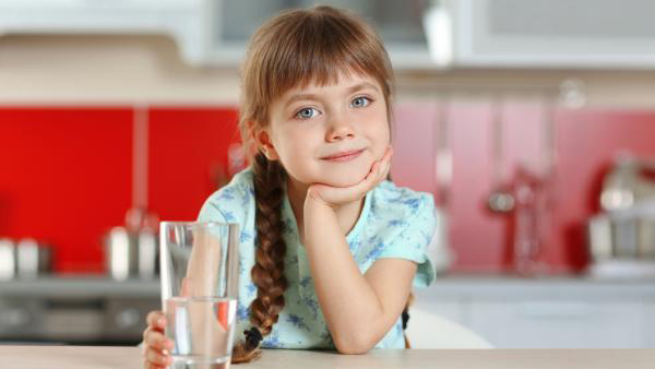 A smiling young schoolgirl in a kitchen holding a glass of water in her right hand