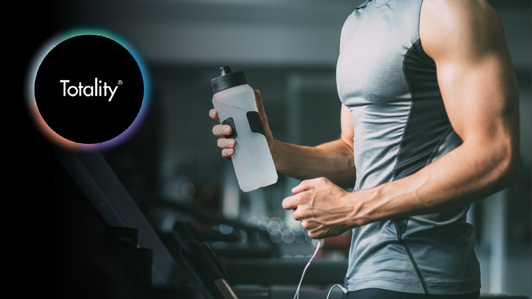 A Totality Logo faintly appearing beside a male athlete running on a treadmill, holding a water bottle A Totality Logo faintly appearing beside a male athlete running on a treadmill, holding a water bottle