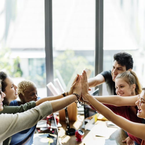 A working office with 6 employees all reaching into a circle to celebrate their collective teamwork