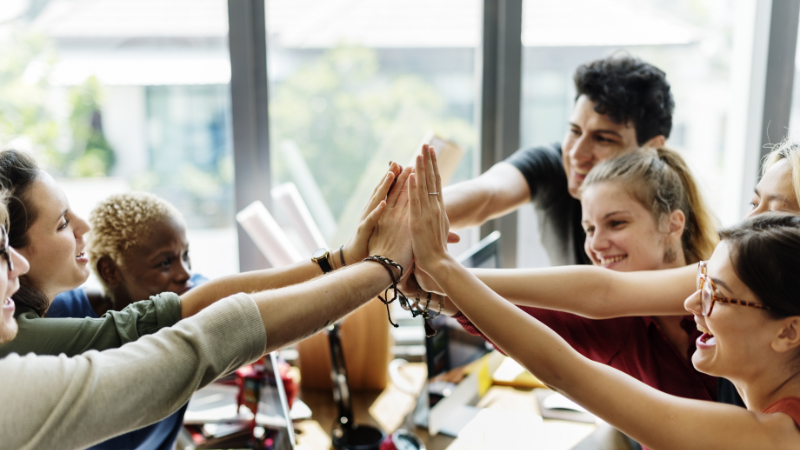 A working office with 6 employees all reaching into a circle to celebrate their collective teamwork