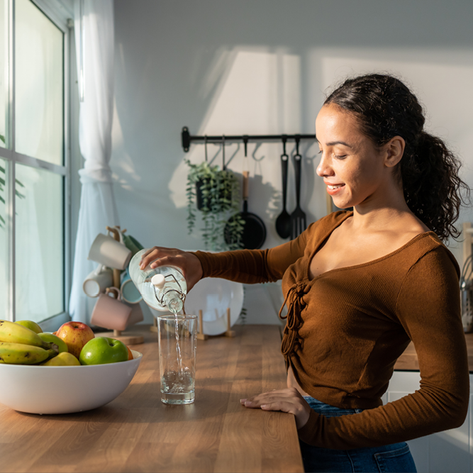 A tanned woman pouring filtered water into a clear glass atop a wooden kitchen countertop