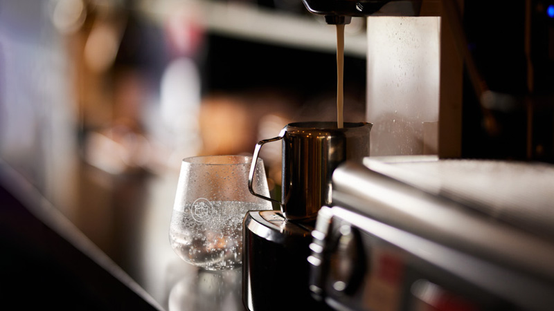 A close-up of a barista bar worktop with a mug of coffee being freshly poured alongside a clear glass of chilled water